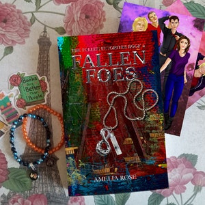May include: A book titled "Fallen Foes: The Fallen Favorites Book 1" by Amelia Rose. The cover features a red, green, and blue abstract background with a silver chain and pendant. The book is surrounded by stickers and bracelets.