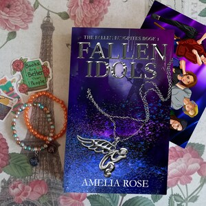 Peut inclure: Une couverture de livre violette et bleue avec le titre "Fallen Idols" et le texte "The Fallen Favorites Book 1" au-dessus du titre. La couverture du livre présente un collier avec une aile d'ange argentée et une image floue d'un groupe sur scène.
