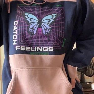 Puede incluir: Una sudadera con capucha azul marino y rosa con un gráfico de una mariposa en un patrón de cuadrícula. El texto "CATCH FEELINGS" está impreso debajo de la mariposa.