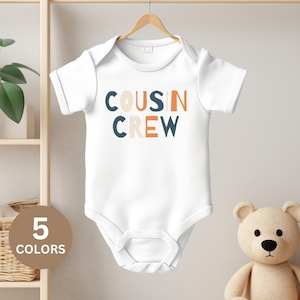 Puede incluir: Un body blanco para bebé con mangas cortas y el texto "COUSIN CREW" en letras de bloque de colores.