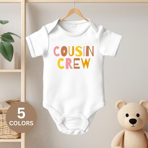 Puede incluir: Un body blanco para bebé con el texto "COUSIN CREW" en letras de colores. Las letras son de color rosa, marrón y amarillo.
