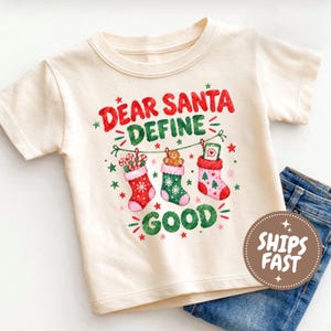 May include: A cream-colored short-sleeve t-shirt featuring a graphic print with the text "DEAR SANTA DEFINE GOOD". The design includes three hanging Christmas stockings, one containing candy canes, one with a gingerbread man, and one with a phone.