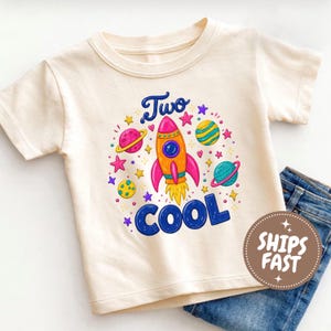 May include: A cream-colored short-sleeve t-shirt featuring a colorful space-themed graphic. The design includes a central orange and pink rocket, planets, and stars, with the blue text Two COOL printed above and below the rocket illustration.