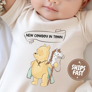 May include: A cream-colored baby bodysuit featuring a graphic of a yellow bear wearing a light blue cape and holding a white spotted hobby horse. Above the bear is a banner with the text NEW COWBOY IN TOWN. A brown circular badge says SHIPS FAST.
