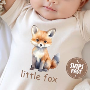 May include: A cream-colored baby onesie featuring a watercolor illustration of a fox. The fox is depicted in shades of orange, brown, and gray, with the words "little fox" printed below. A circular sticker with the words "SHIPS FAST" is also visible.