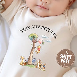May include: A cream-colored baby bodysuit featuring a graphic illustration of Winnie the Pooh characters under a tree with a balloon. The text TINY ADVENTURER is printed in black above the illustration. A brown circular badge with white text is visible.
