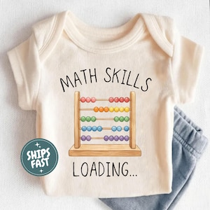 May include: A cream-coloured baby bodysuit featuring a graphic of a wooden abacus with colourful beads. The text "MATH SKILLS" is printed in black above the abacus, and "LOADING..." is printed in black below it. A circular teal badge reads "SHIPS FAST".