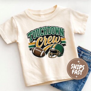Könnte beinhalten: Ein cremefarbenes Kurzarm-T-Shirt mit einem Grafikdruck, der den Text TOUCHDOWN Crew in Grün und Gold zeigt. Das Design umfasst einen braunen Football und einen grünen Football-Helm mit goldenen Akzenten vor einem abgenutzten Hintergrund.