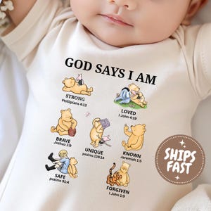 Può includere: Un body per neonati color crema con il testo GOD SAYS I AM e illustrazioni dei personaggi di Winnie the Pooh. La grafica include etichette come STRONG, LOVED, BRAVE, UNIQUE, KNOWN, SAFE e FORGIVEN, ognuna abbinata a un versetto biblico.