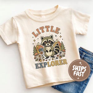 May include: A cream-coloured short-sleeve t-shirt featuring a graphic of a raccoon standing in a forest with mountains, mushrooms, and flowers. The text "LITTLE EXPLORER" is printed in a curved font above and below the raccoon illustration.