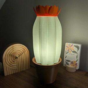 May include: A white and orange ceramic lamp shaped like a cactus with a green light shining from inside. The lamp is sitting on a wooden table with a wooden clock and a calendar.