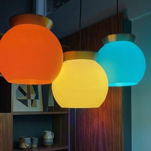 May include: Three pendant lights with spherical shades in orange, yellow, and blue. Each light has a gold-colored top. The shades have a textured, ribbed appearance. The lights hang in front of a wooden cabinet.