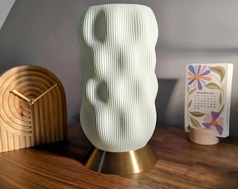May include: A white ceramic lamp with a textured, wavy design and a gold base. The lamp is sitting on a wooden surface.
