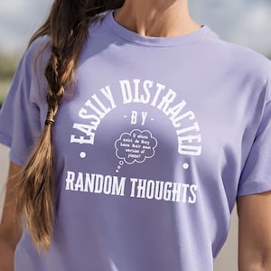 以下が含まれることがあります： ラベンダー色のTシャツに、白い文字で「Easily Distracted by Random Thoughts」と書いてあります。テキストは円の中にあり、思考の泡の中に「もしエイリアンが存在するなら、彼らは自分たちのピザバージョンを持っているのだろうか？」と書かれています。