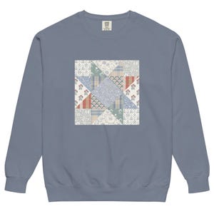 May include: A dusty blue sweatshirt featuring a patchwork design. The square patch is composed of various patterned fabrics in shades of blue, red, and cream. The sweatshirt has a crew neck and long sleeves.