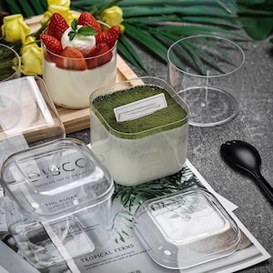 May include: Clear plastic dessert cups and containers filled with layered desserts. One cup has strawberries and cream, another has a green powder topping. The image also shows a black spoon and a clear glass.