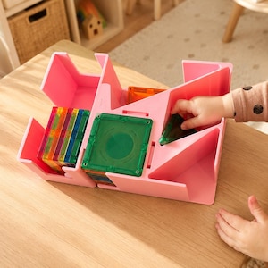 May include: A pink plastic storage organizer with multiple compartments designed for magnetic tiles. The organizer holds several colorful translucent square and triangular magnetic tiles in various shades of red, orange, yellow, green, and blue.