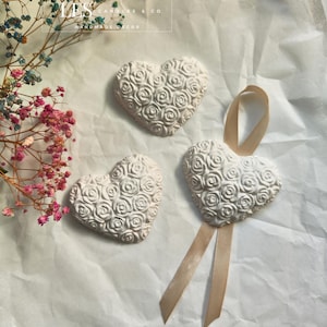 Op de afbeelding: Drie witte keramische hartvormige ornamenten met een reliëf van rozen. Een hart heeft een beige lint.