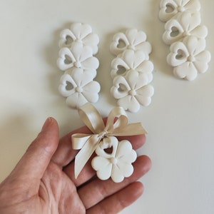 Op de afbeelding: Witte keramische bloemencharms met een hartvormig uitsnijding. De charms zijn gestapeld in groepen van drie en gebonden met een beige lint.