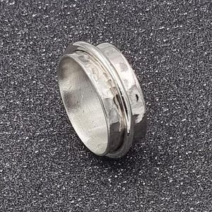 Könnte beinhalten: Ein silberfarbener Spinner-Ring mit einer gehämmerten Textur auf dem äußeren Band. Der Ring hat ein zentrales Band, das sich frei dreht. Der Ring ist vor einem dunklen, strukturierten Hintergrund platziert.