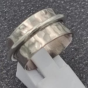 Könnte beinhalten: Ein silberfarbener Ring mit gehämmerten Texturen auf dem Band. Der Ring hat ein sich drehendes äußeres Band. Der Ring wird auf einem weißen Ständer vor einem dunklen, strukturierten Hintergrund präsentiert.