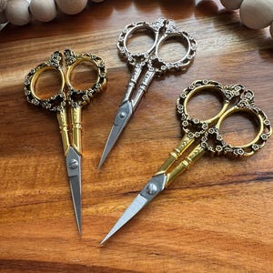 May include: Three pairs of decorative scissors with ornate floral designs. The scissors are gold and silver in color and have a vintage aesthetic.