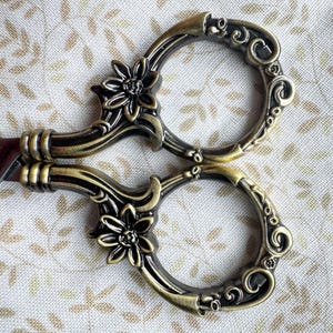 May include: Ornate gold-toned scissors with floral details. The scissors have a decorative design with intricate scrollwork and flower motifs.