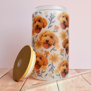 May include: A glass can-shaped tumbler with a bamboo lid and a clear straw. The tumbler is decorated with a pattern of golden doodle dog faces and yellow flowers on a white background. The lid is made of bamboo.