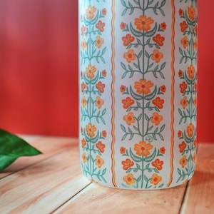Pu&ograve; includere: Vaso in ceramica bianca con un motivo floreale in arancio e verde. Il vaso ha una forma cilindrica e una finitura liscia e lucida.