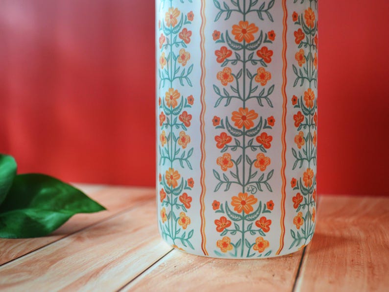 Pu&ograve; includere: Vaso in ceramica bianca con un motivo floreale in arancio e verde. Il vaso ha una forma cilindrica e una finitura liscia e lucida.
