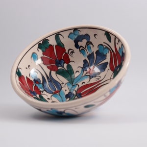 Könnte beinhalten: Eine cremefarbene Keramikschale mit floralem Muster. Die Blumen sind in Rot, Blau und Grün mit schwarzen Akzenten bemalt. Die Schale hat eine leicht abgewinkelte Form und eine glatte, glänzende Oberfläche.