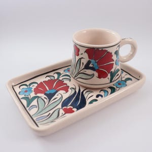 May include: A white ceramic coffee cup and saucer set featuring a traditional Turkish floral design. The cup and saucer are decorated with red, blue and green flowers and leaves.