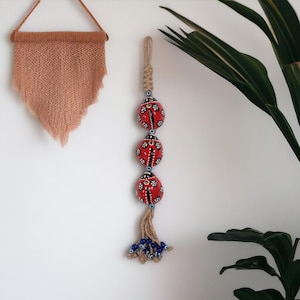 Peut inclure: Trois ornements en céramique rouge en forme de coccinelle avec des détails blancs et noirs sont suspendus à une corde de jute avec des perles bleues en forme d'œil maléfique. Les ornements sont enfilés en ligne verticale et ont un pompon en bas.