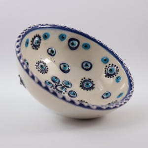Può includere: Una ciotola in ceramica color crema con bordo blu, decorata con disegni a forma di occhio blu e bianco. Il bordo ha un bordo smerlato con un bordo blu. La ciotola è su uno sfondo bianco.