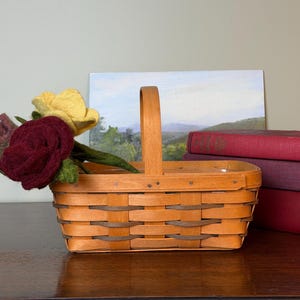 May include: A rectangular woven wood basket with a single upright handle, featuring light brown horizontal slats and darker brown accent strips. Inside the basket are several felt flowers in shades of deep burgundy and muted yellow.