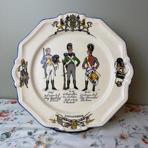 May include: A cream-colored ceramic plate with a scalloped edge and blue trim, featuring three historical infantry soldiers in uniform, a coat of arms at the top, and military equipment illustrations. Text includes 1807, 1812, 1806, and INFANTERIE.