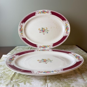 May include: Two oval ceramic serving platters featuring a cream-colored base with a wide, deep red border. The rim is decorated with colorful floral clusters in yellow, pink, and blue, with a matching floral bouquet centered on each platter.