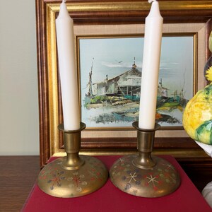 May include: Two brass candle holders with wide, circular bases featuring a colorful starburst pattern in red, blue, and green. Each holder supports a single, plain white taper candle. The metal has a warm, aged finish with visible patina.