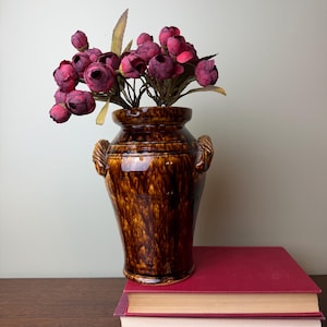 May include: A glossy brown ceramic vase with two side handles, featuring a mottled glaze finish. The vase is filled with a bouquet of dark pink artificial flowers with green leaves, resting atop two stacked red hardcover books.