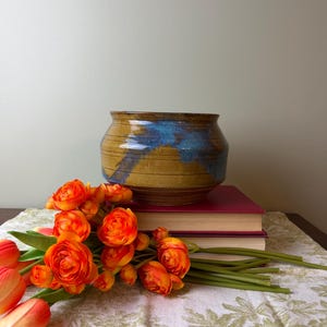 May include: A ceramic bowl with a wide mouth and a tapered base, featuring a mustard yellow glaze with a prominent blue drip pattern. The bowl has horizontal incised lines around its body and a smooth, glossy finish on the exterior surface.