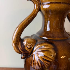 May include: A glossy brown ceramic vase featuring a crackle glaze finish. The vessel is adorned with sculpted elephant heads on the sides, where the trunks extend upward to form the handles, connecting to the neck of the vase.