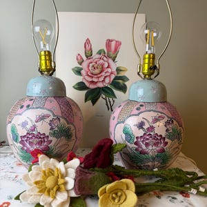 May include: Two matching ceramic ginger jar lamps with a light blue lid and a pink body featuring painted lotus flowers and green leaves. Each lamp has a brass-toned metal harp and a clear light bulb. Felt flowers in white, yellow, and red sit in front.