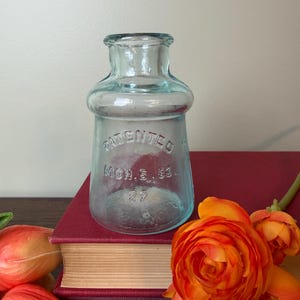 May include: A clear, light blue glass bottle with a wide neck and a rounded shoulder. The front of the bottle features embossed text that reads "PATENTED MCH. 5. 80." with the number "27" below it, indicating a vintage glass container.
