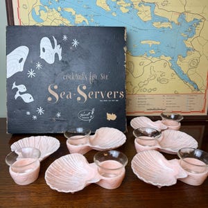 May include: A set of six pink shell-shaped snack dishes with integrated clear glass cups, presented with a black box labeled "cocktails for six" and "Sea-Servers". The dishes feature a ridged texture and are arranged in front of the box.