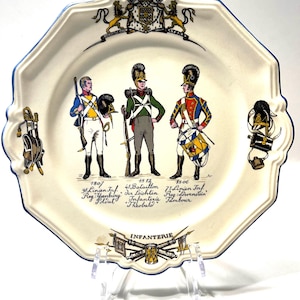 May include: A white ceramic plate with a blue rim features a depiction of three soldiers in 19th-century military uniforms. The plate includes the text "INFANTERIE" and the dates "1807", "1812", and "1806".