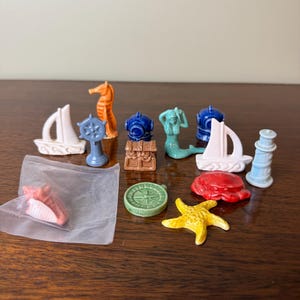 Peut inclure: Une collection de petites figurines en céramique sur le thème nautique comprenant deux voiliers blancs, un hippocampe orange, une barre de navire bleue, deux casques de plongée bleus, un coffre au trésor marron, une sirène, un phare bleu clair, une boussole verte, un crabe rouge, une étoile de mer jaune et un coquillage rose dans un sac en plastique.