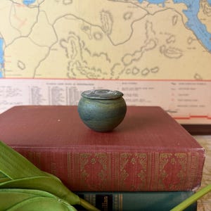 May include: A small, round, lidded ceramic jar with a textured, mottled green and dark blue finish. The lid features a circular, etched design in a lighter, muted tone. The jar has a bulbous shape and sits as a singular, compact object.
