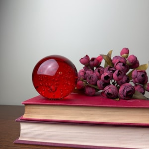 May include: A spherical red glass paperweight containing numerous small air bubbles, positioned on top of two stacked hardcover books with red covers, accompanied by a cluster of small, dark pink artificial rosebuds with green leaves.