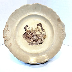 May include: A vintage ceramic plate with a brown and white image of the Duke and Duchess of Edinburgh. The plate has a scalloped edge and a floral design.