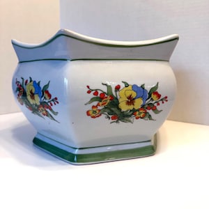 May include: White ceramic planter with a green trim and floral design. The planter features a square base and a curved top edge. The floral design includes yellow, blue, and red flowers with green leaves.